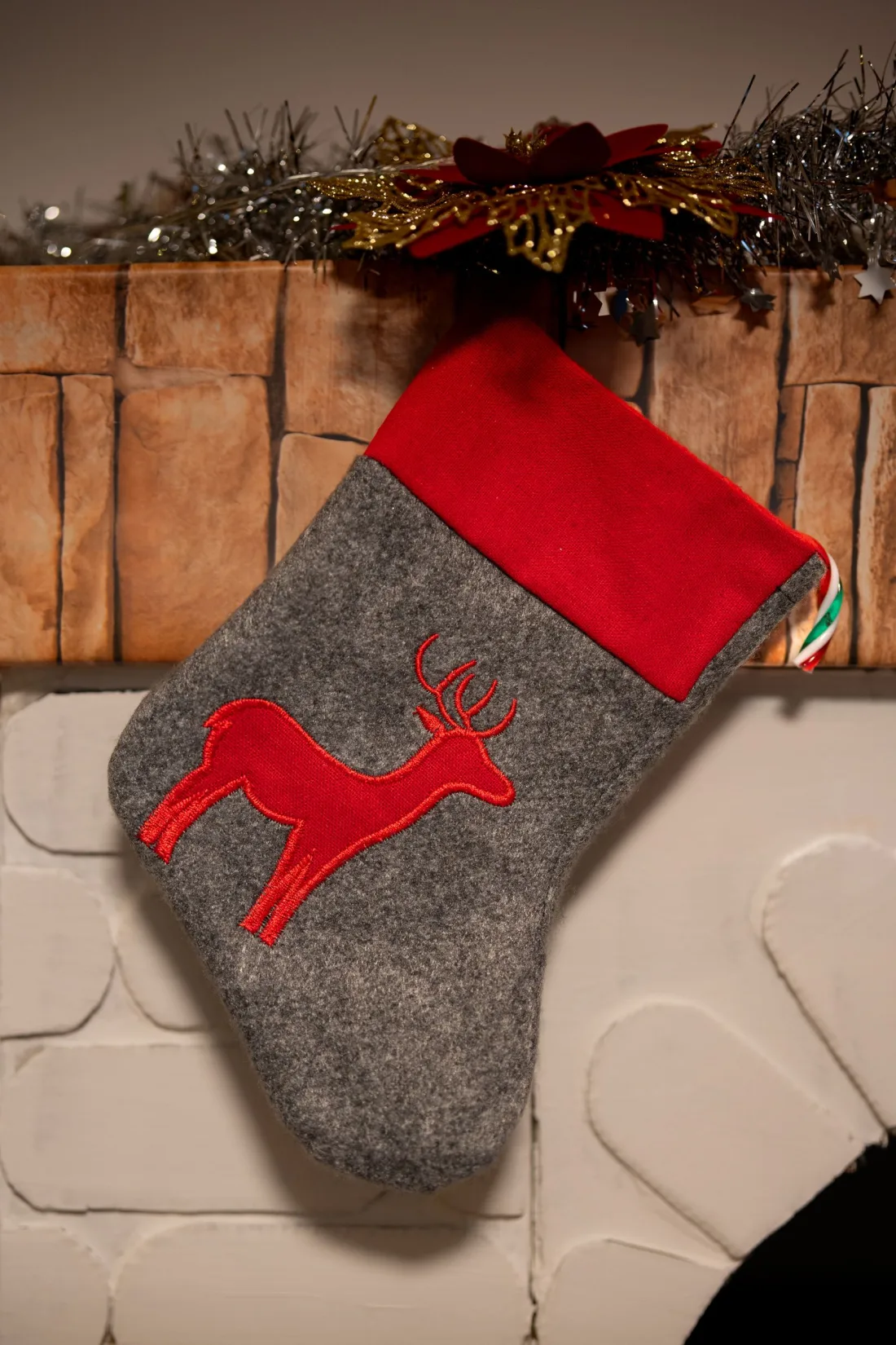 A gray felt Christmas stocking with a red cuff and a red felt reindeer stitched on, hanging from a mantel
