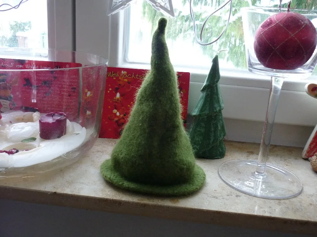 A cone-shaped Christmas tree knitted in green wool and then felted, standing on a windowsill beside a smaller tree