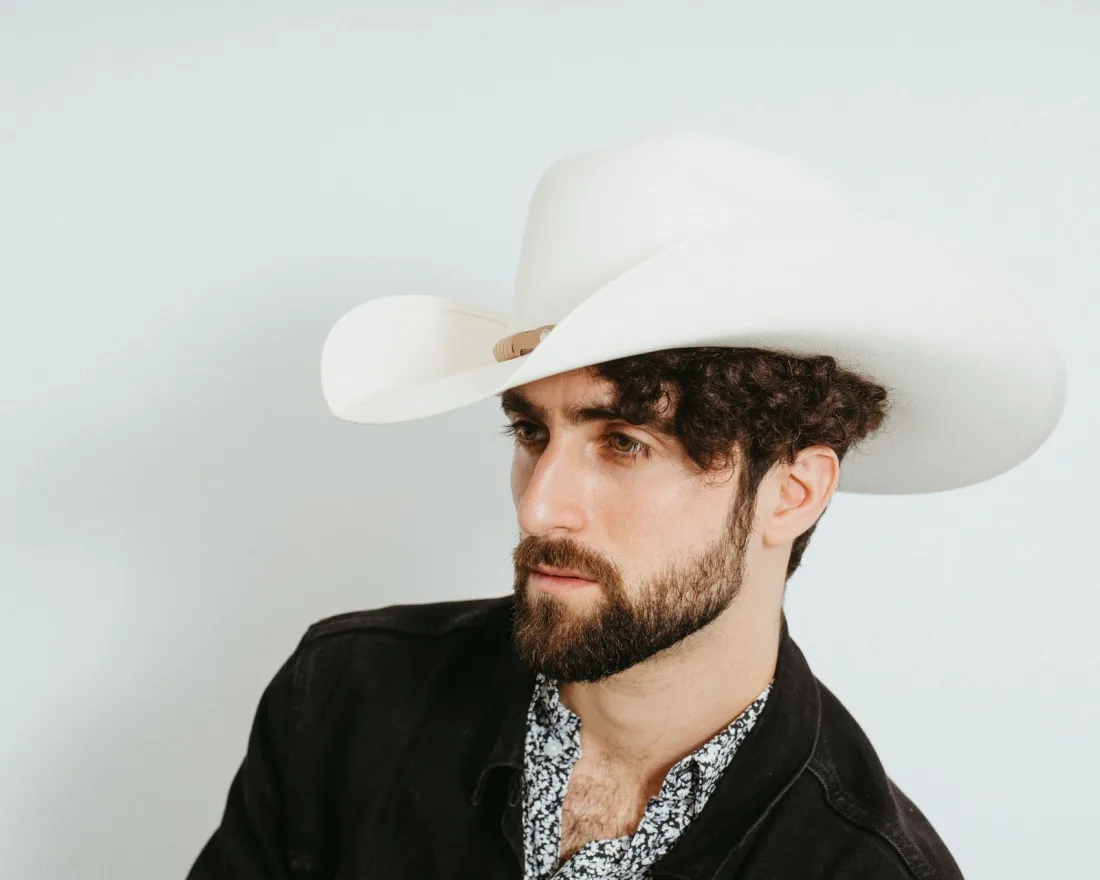 A bearded man wearing a cream felt cowboy hat with a tall crown and a wide brim curled up at the sides