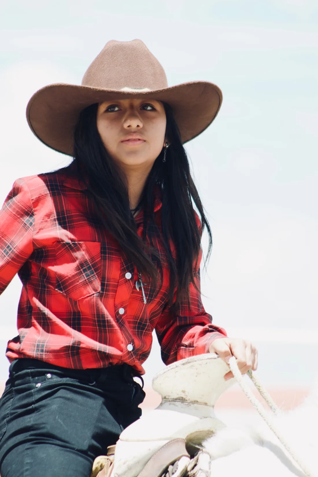 A woman with long dark hair worn loose under a taupe felt cowboy hat, in a red plaid shirt, holding a rope at a white saddle