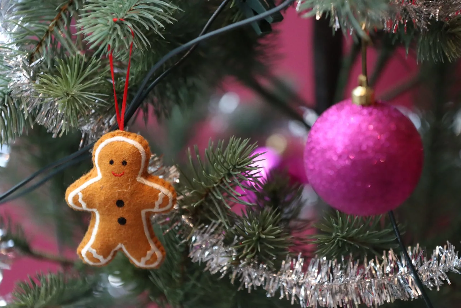 A stuffed golden-brown felt gingerbread man ornament with a wavy white stitched icing outline, black dot eyes, a red stitched smile and two black buttons, hanging on a red ribbon loop beside a pink bauble