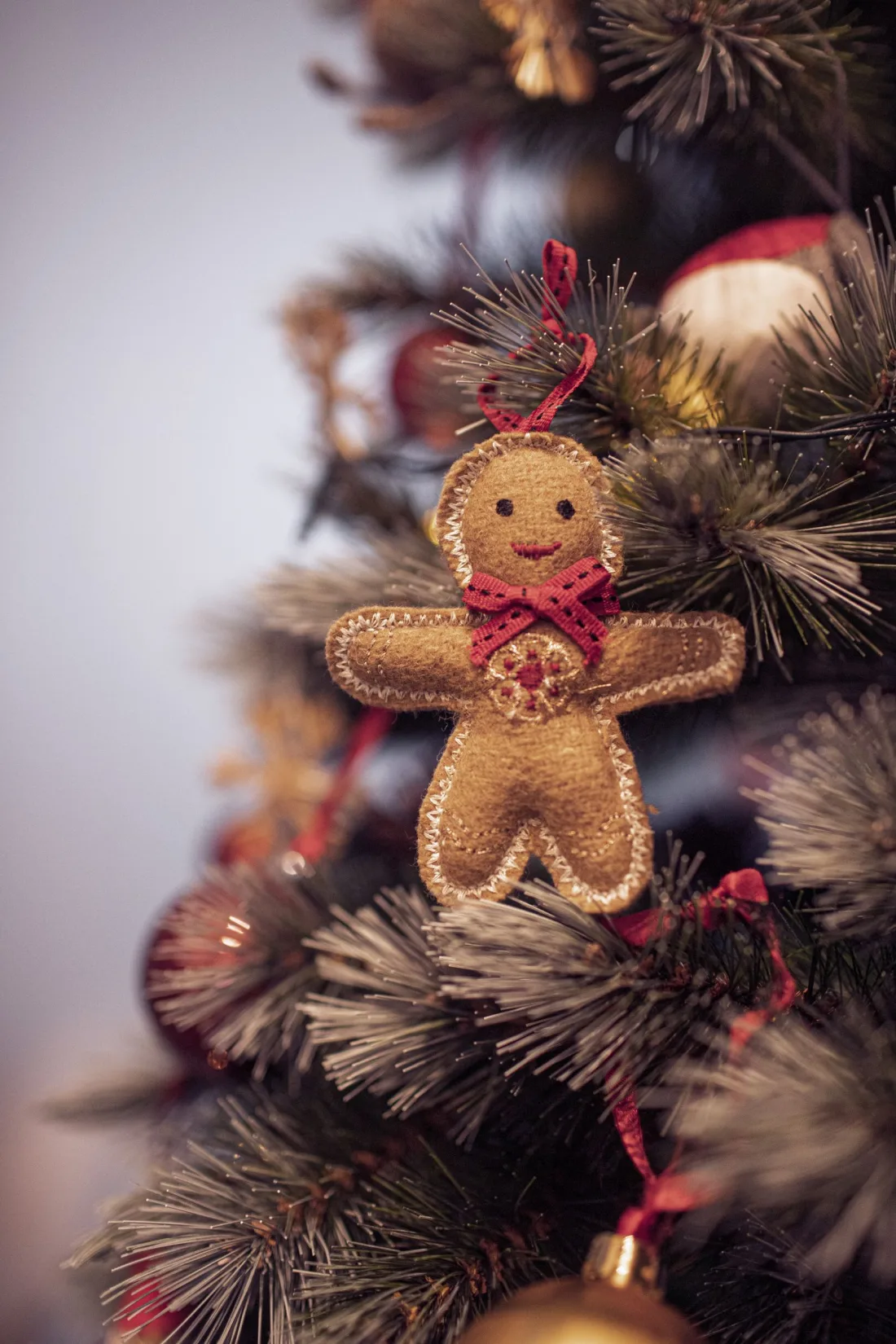 A tan felt gingerbread man ornament edged with white zigzag stitching, with an embroidered face, a red ribbon bow and a stitched red and gold medallion on its chest, on a frosted Christmas tree
