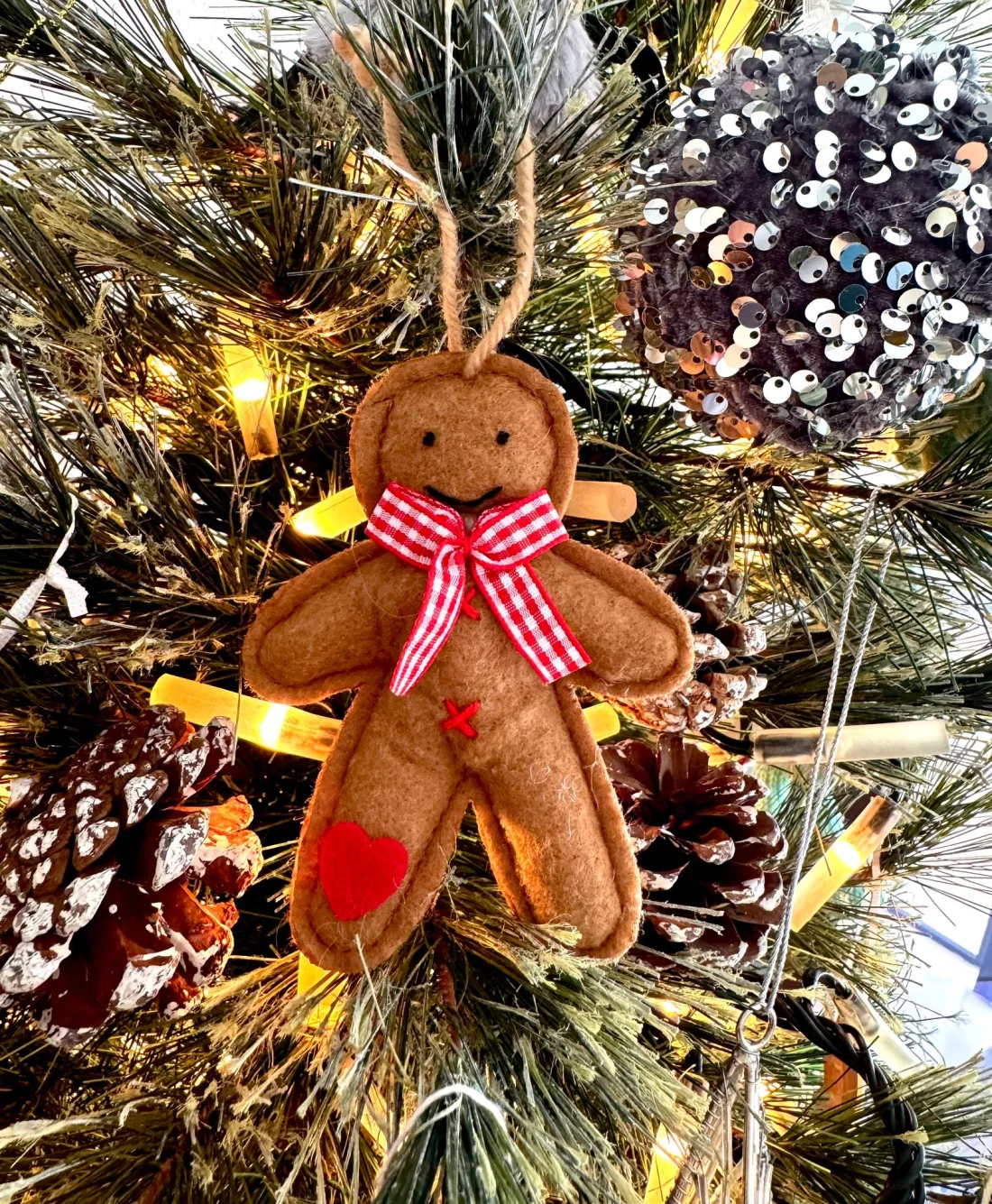 A brown felt gingerbread man ornament with a stitched black face, a red gingham bow, red cross-stitch buttons and a red felt heart on one leg, hanging from a twine loop on a Christmas tree