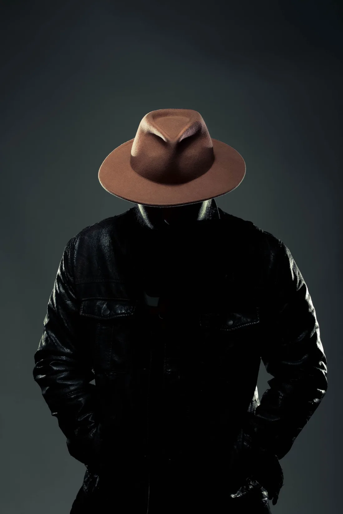 A man in a black leather jacket wearing a brown felt fedora with a pinched front and a narrow tonal band, his face hidden under the brim