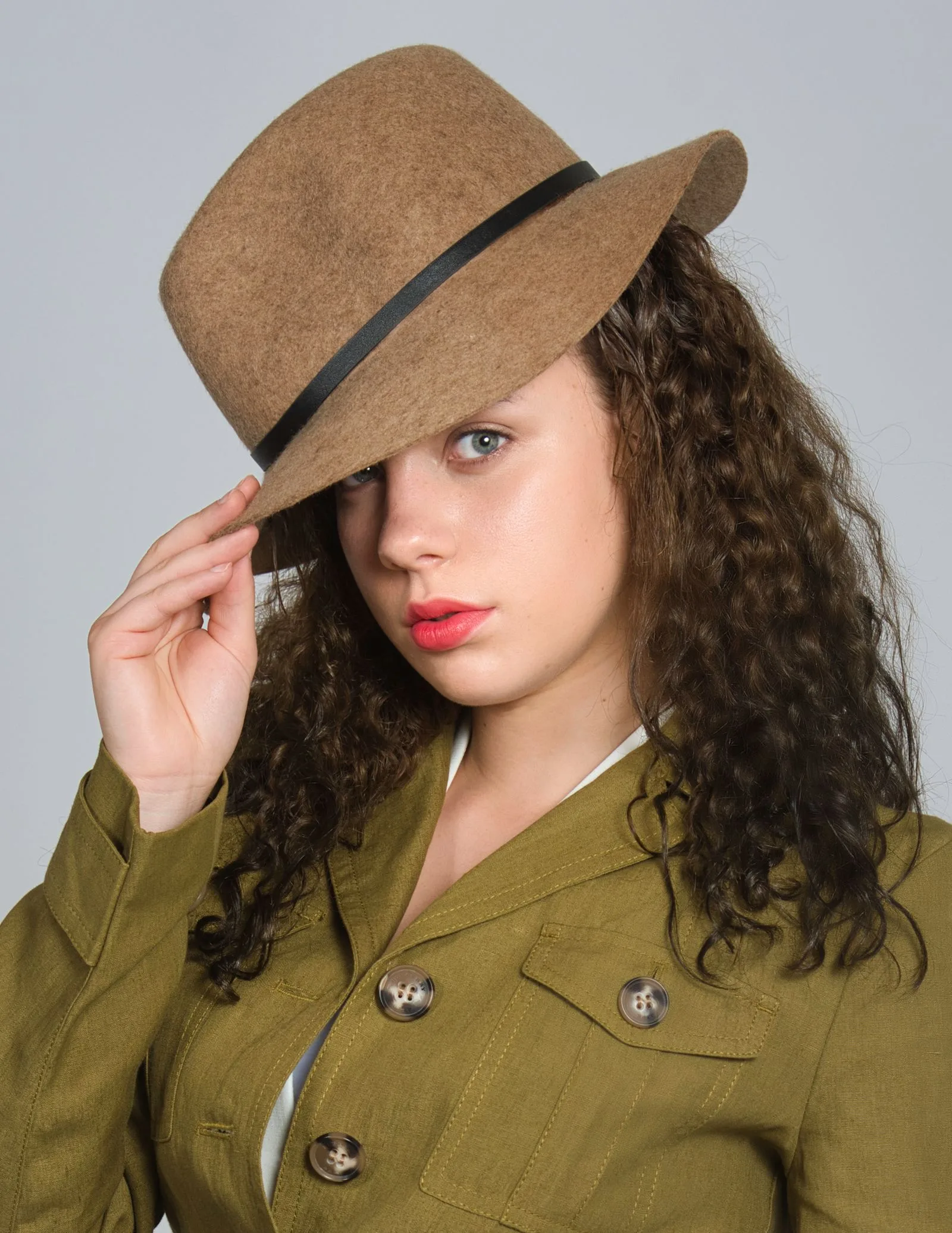 A woman with long curly dark hair touching the brim of a camel felt fedora with a thin black band
