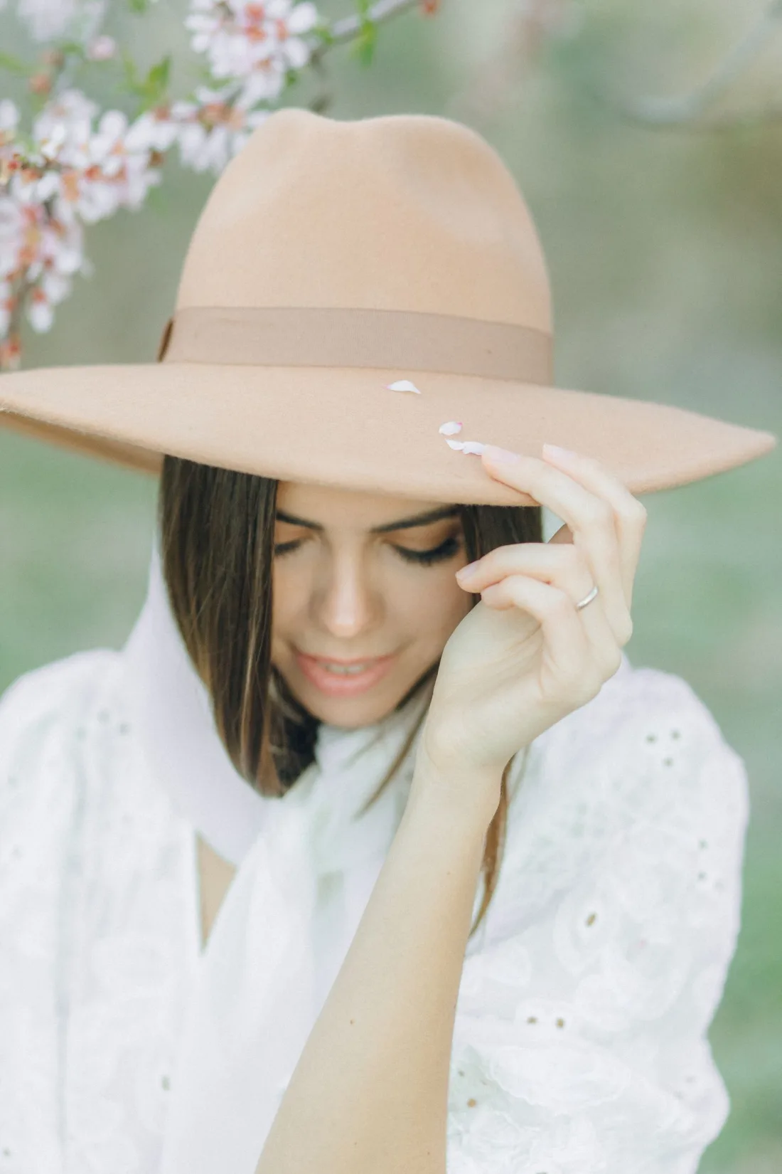 A woman in a white blouse holding the flat brim of a wide beige felt hat with a tonal band, blossom behind her