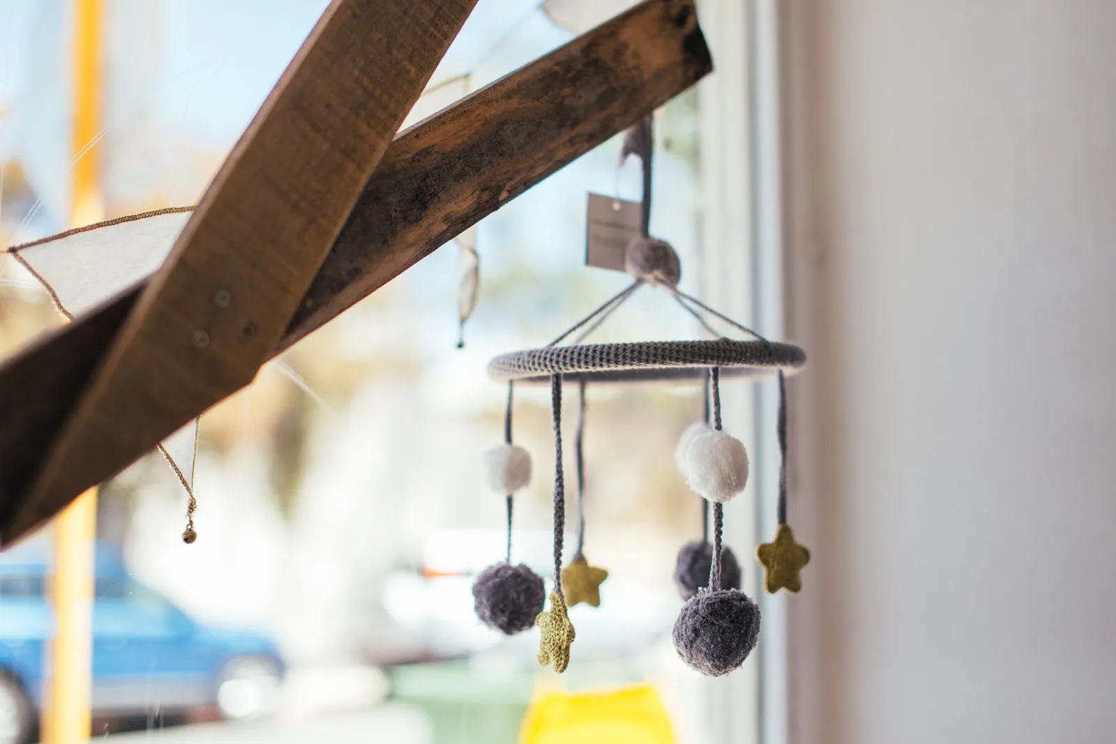 A hoop mobile hung with strands of gray and white wool balls and small stars, hanging from a wooden beam beside a window