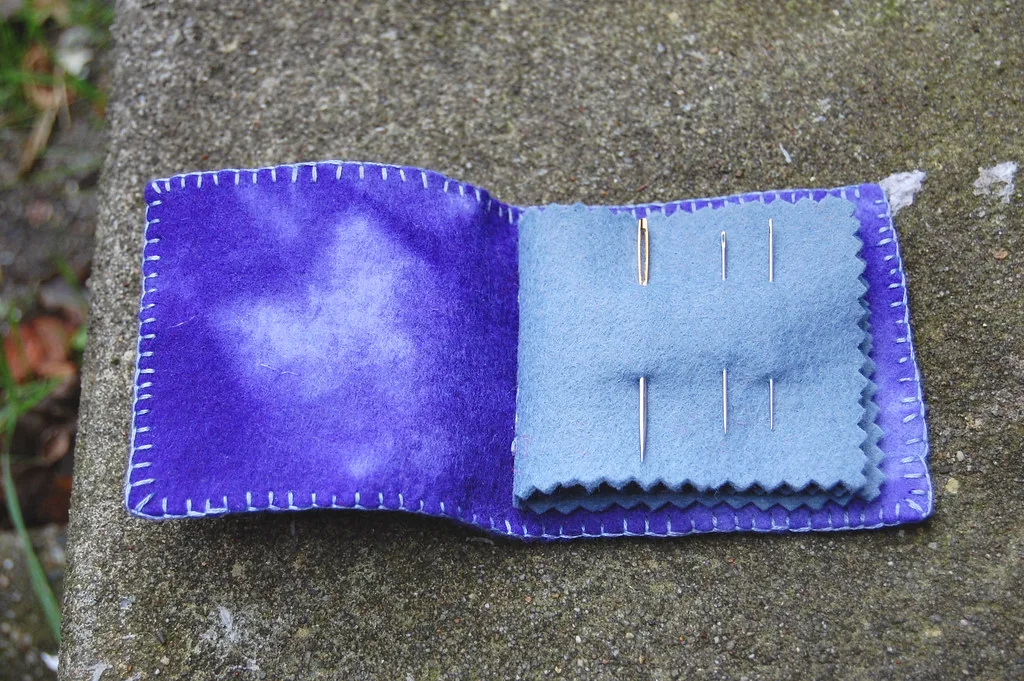 An open purple felt needle book with blanket-stitched edges and a stack of light blue felt pages cut with pinked zigzag edges, three sewing needles woven into the top page