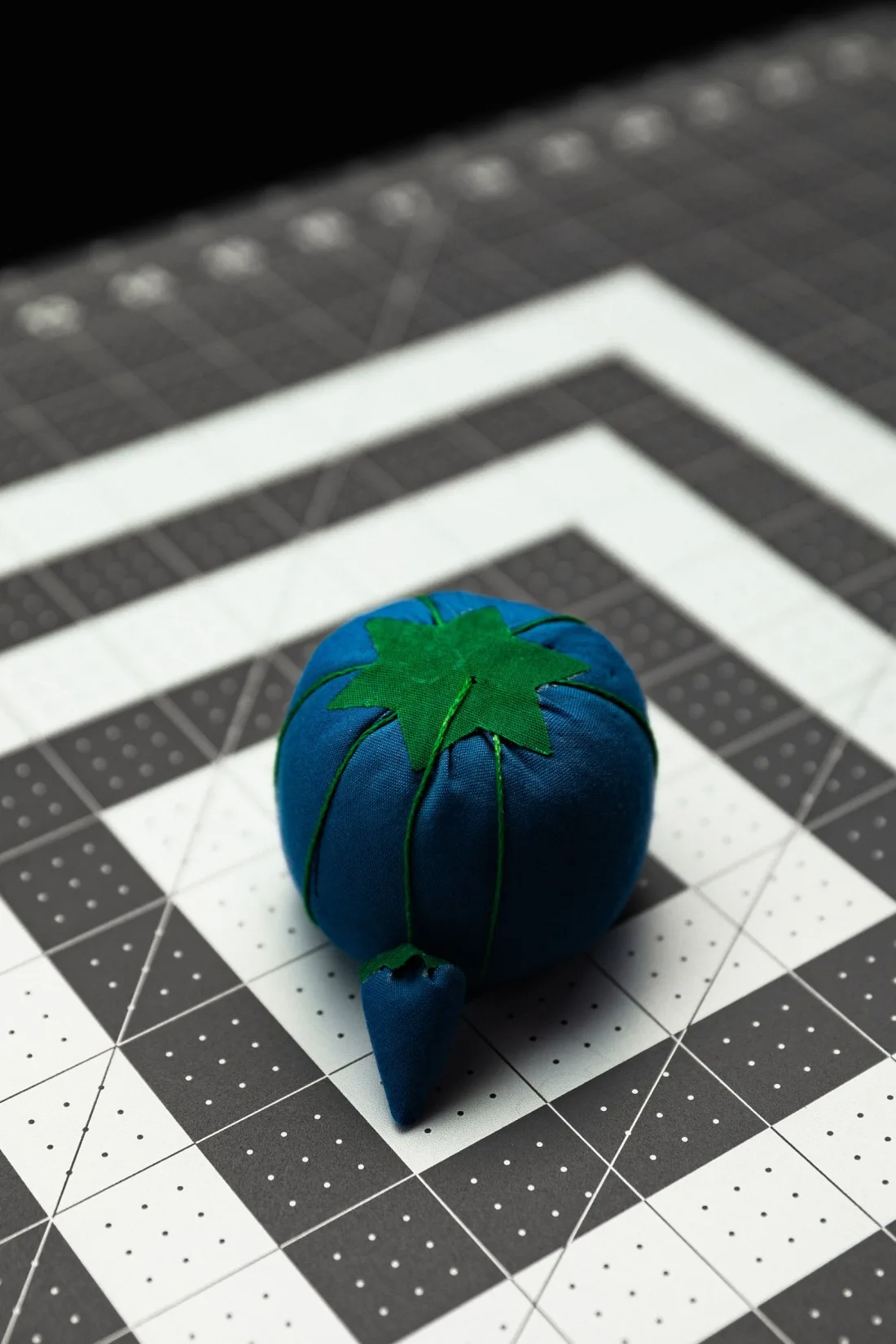 A blue fabric tomato-style pincushion with a green star-shaped top, green thread wraps dividing it into lobes and a small attached strawberry, on a grey cutting mat