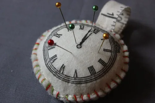 A round felt pincushion printed with a clock face, with coloured glass-head pins standing in it and a coiled tape measure behind
