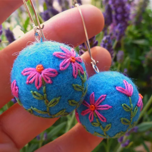 A pair of turquoise felted ball earrings embroidered with pink flowers and green leaves, held up in front of lavender