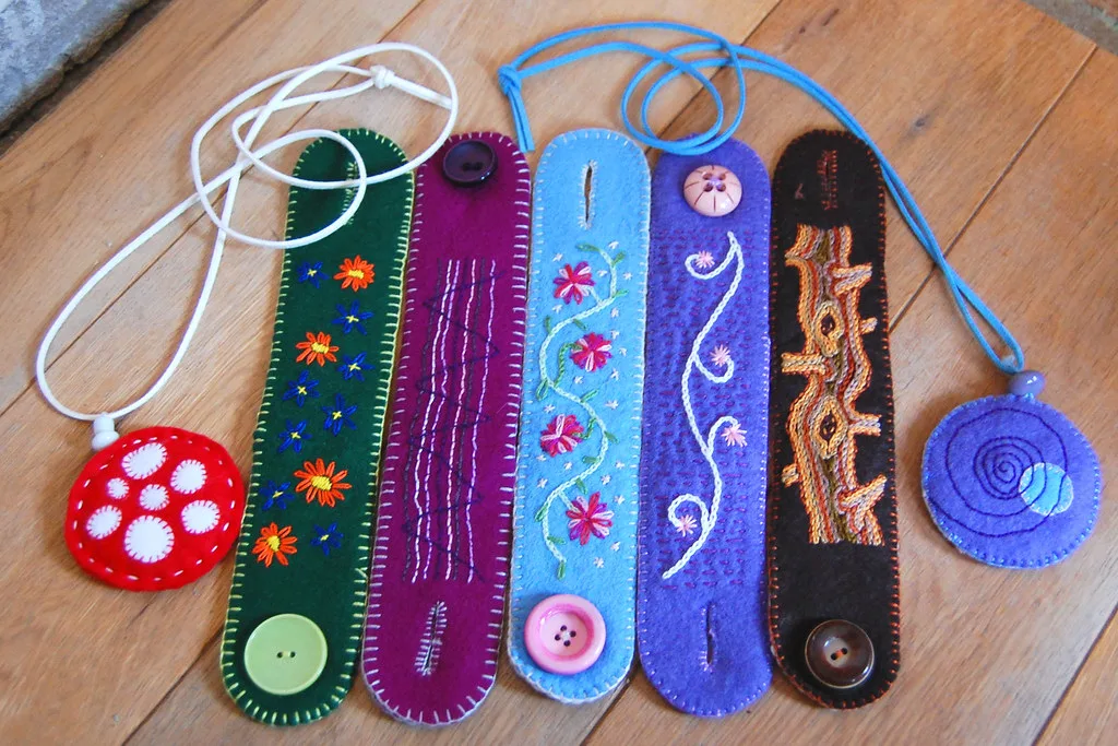 Five felt cuff bracelets with embroidered flowers and swirls and button closures, beside two round felt pendants on cords