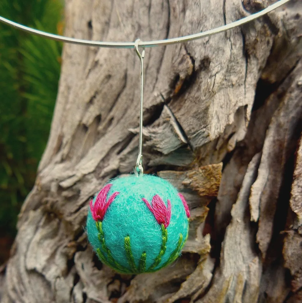A turquoise felted ball pendant embroidered with pink tulips, hanging from a silver neck wire