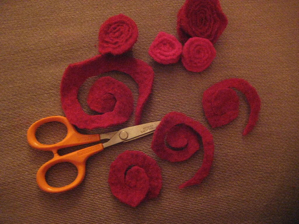 Red spirals cut from felted sweater wool beside orange scissors, some already rolled into roses