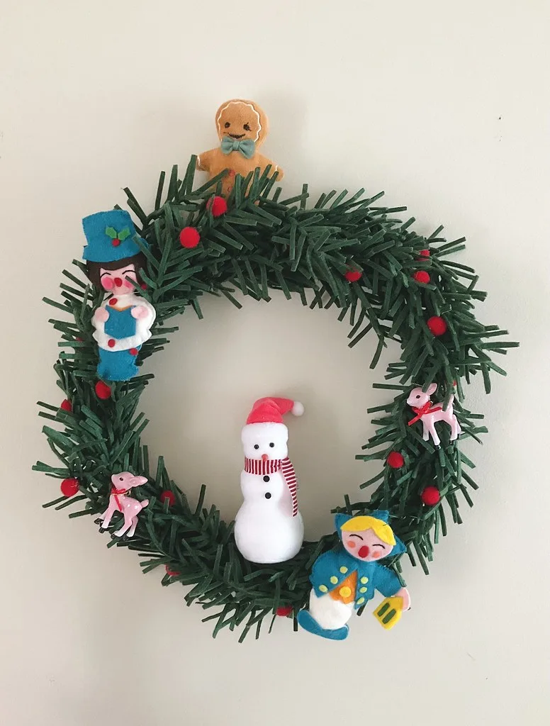 A green fringed Christmas wreath with small figures, a snowman and red berries