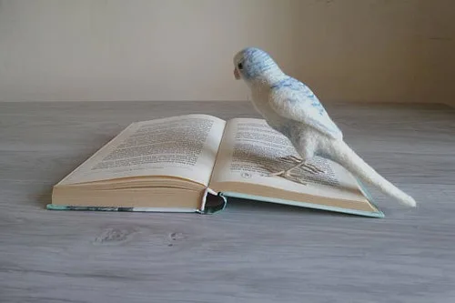 A pale blue needle felted budgie with a long tail standing on the pages of an open book