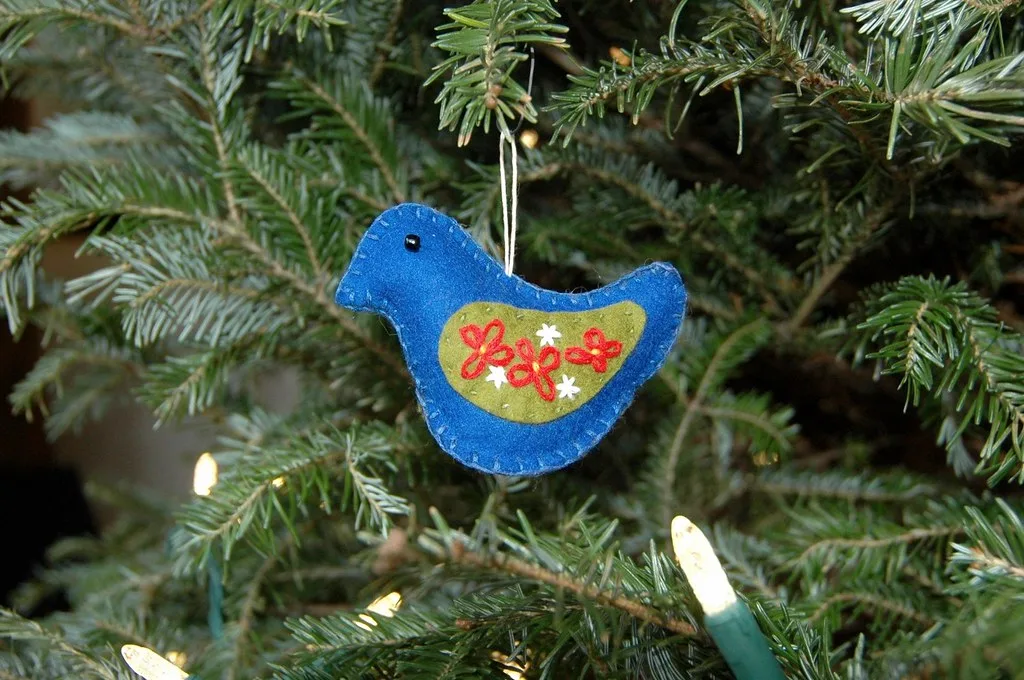 A blue sheet felt bird ornament with an embroidered green wing panel hanging on a lit Christmas tree