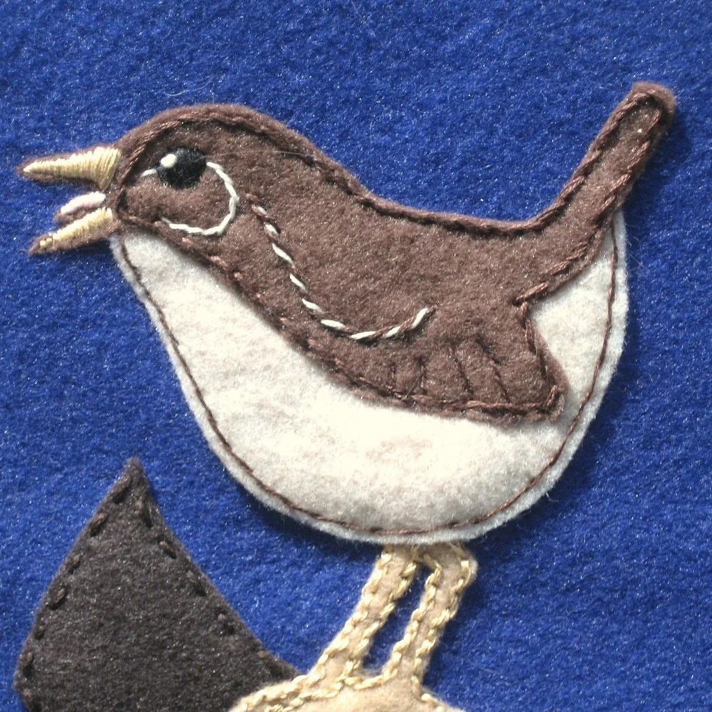 A felt wren appliquéd onto blue felt, in brown and cream with stitched wing lines and a cocked tail