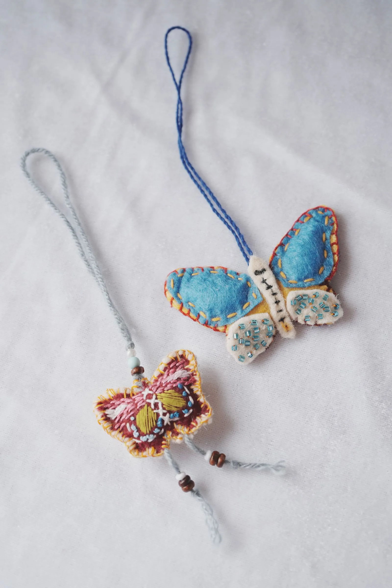 Two handmade felt butterfly ornaments on cord loops: a blue one with cream hind wings sewn with turquoise beads, and a smaller red and pink one covered in embroidery