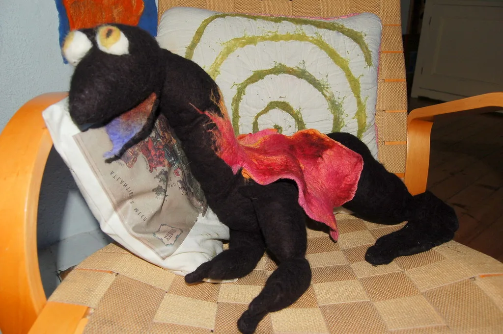 A large black felted dragon with long legs and pale eyes lying across a cushion on a chair, its red and pink felt wings spread over its back