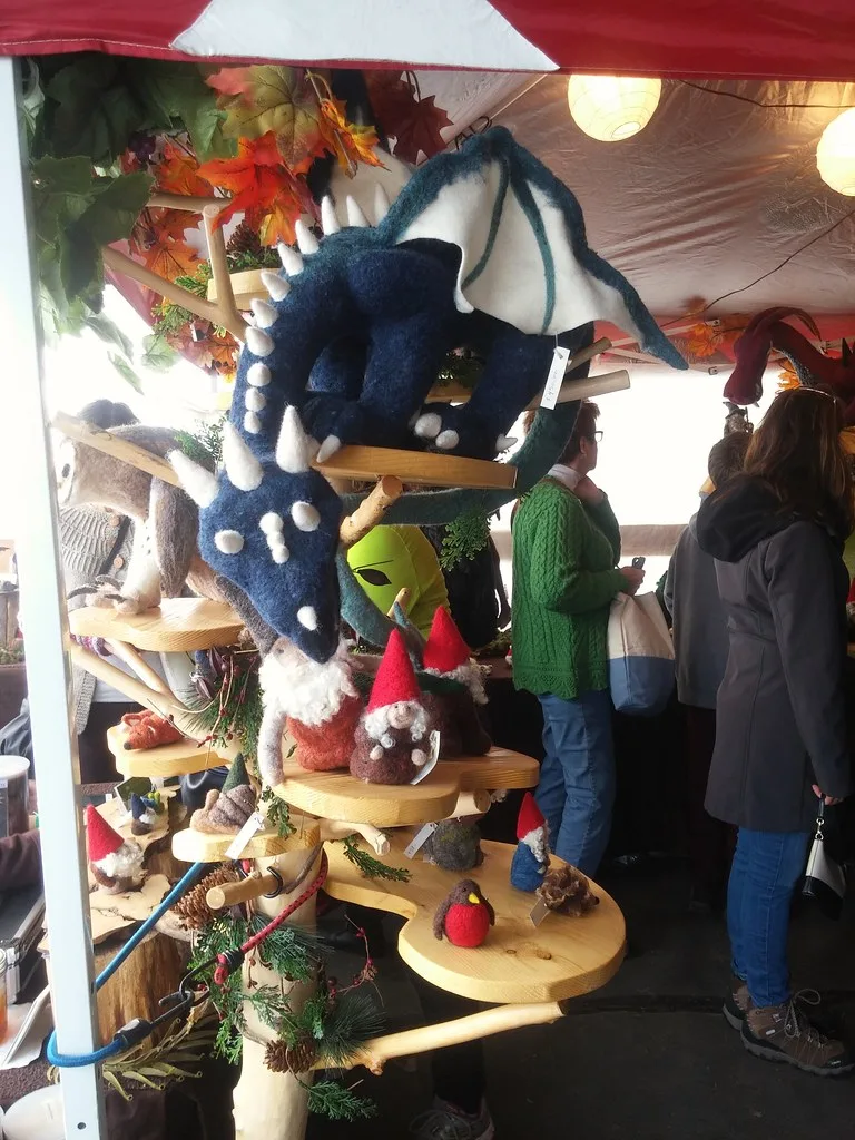 A large dark blue felted dragon with white spikes along its back and white-spotted sides, draped over a wooden display stand above felted gnomes at a craft fair, its blue-veined white wing raised
