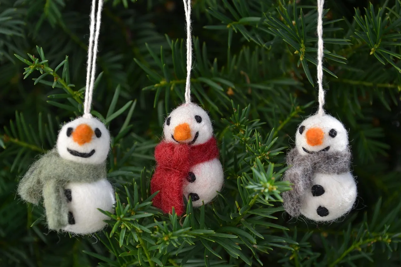 Three needle felted snowman ornaments with orange noses and fringed wool scarves hanging on a fir tree