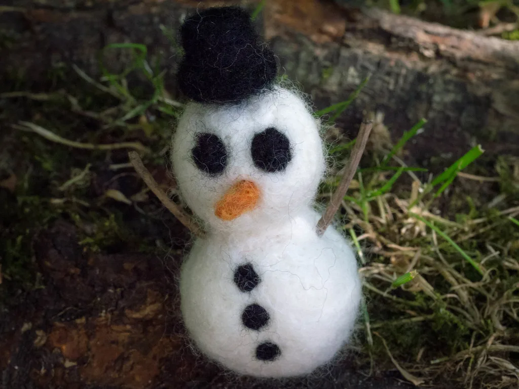 A two-ball needle felted snowman with a black top hat, twig arms, an orange nose and black buttons